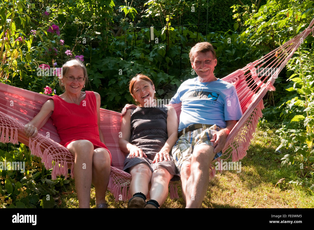 Three very happy looking middle-aged people, couple and friend, sitting on a hammock in afternoon sunshine Stock Photo