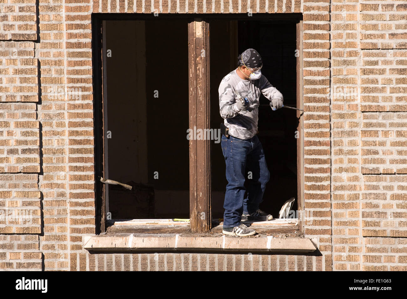 Construction Worker w/ a large chisel and hammer removing & replacing a wooden window frame from a brick building Stock Photo