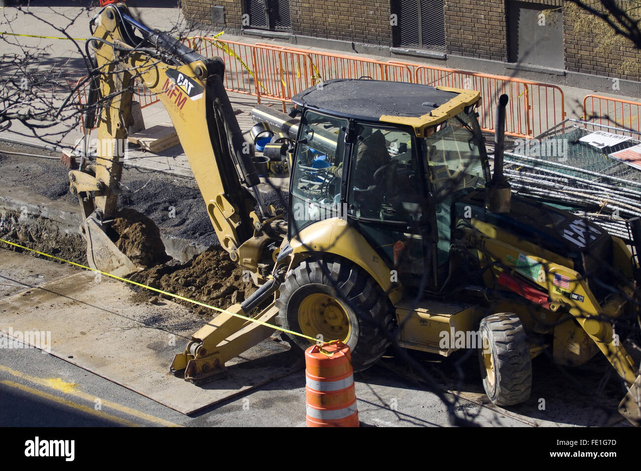 Construction Worker operating a yellow Backhoe made by the Caterpillar Corporation that is digging up a city street Stock Photo