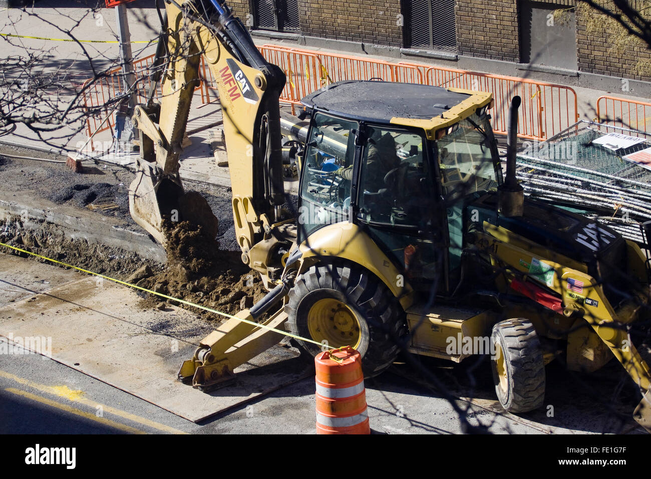 Construction Worker operating a yellow Backhoe made by the Caterpillar Corporation that is digging up a city street Stock Photo