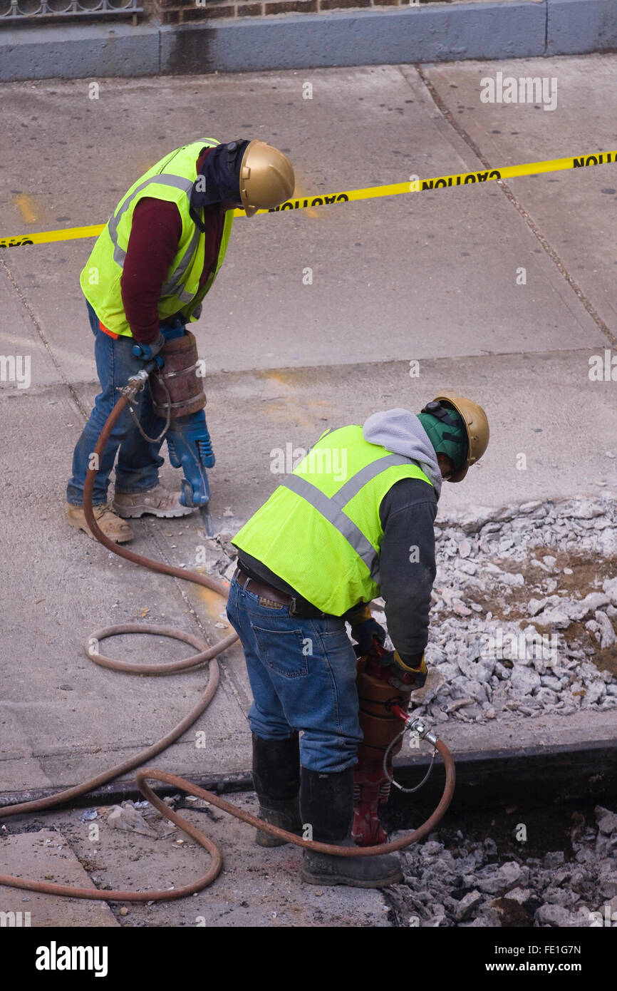 Two Construction Workers with Jackhammers break up the concrete of a city street and sidewalk Stock Photo