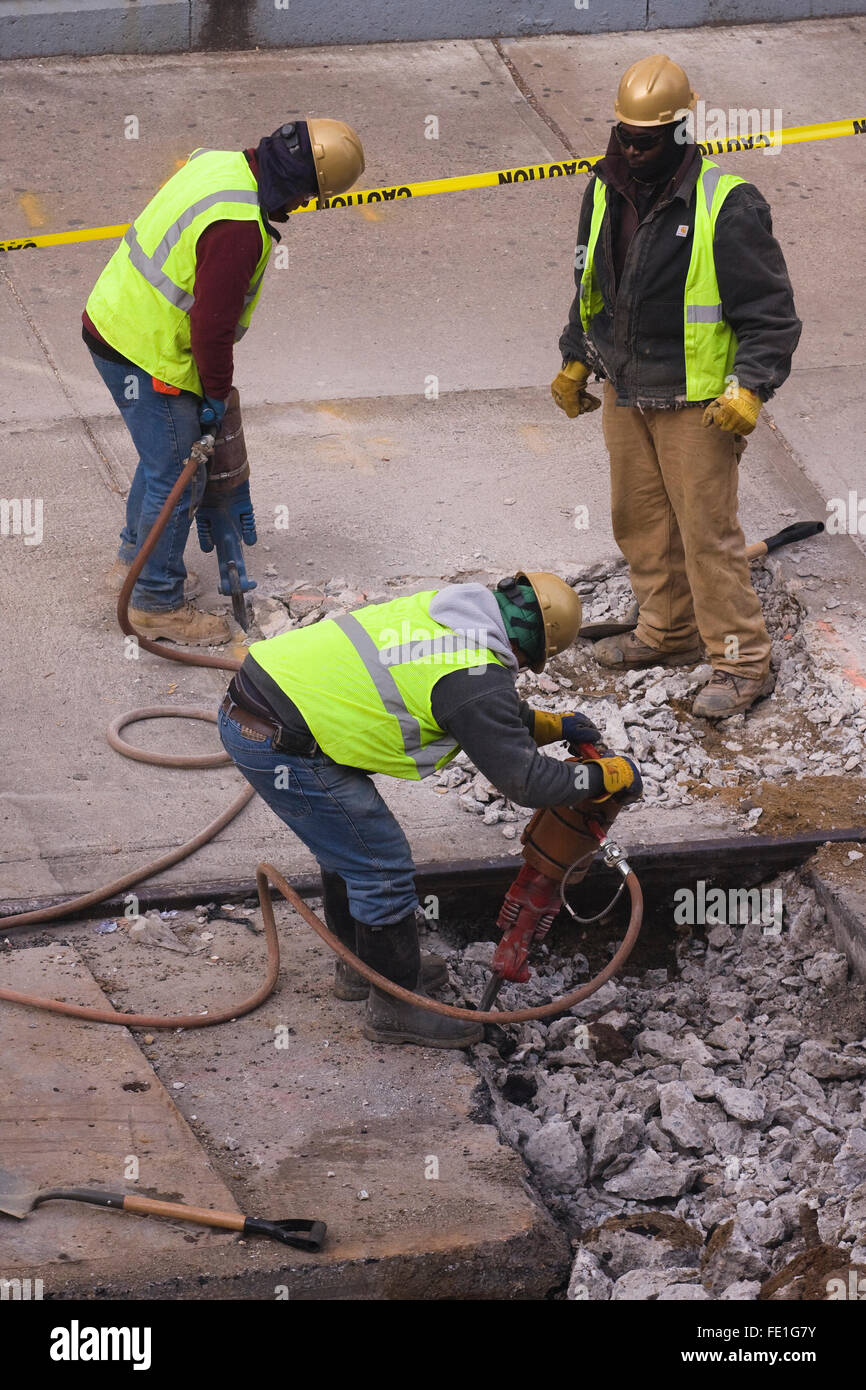 Three Construction Workers two with Jackhammers break up the concrete of a city street and sidewalk Stock Photo