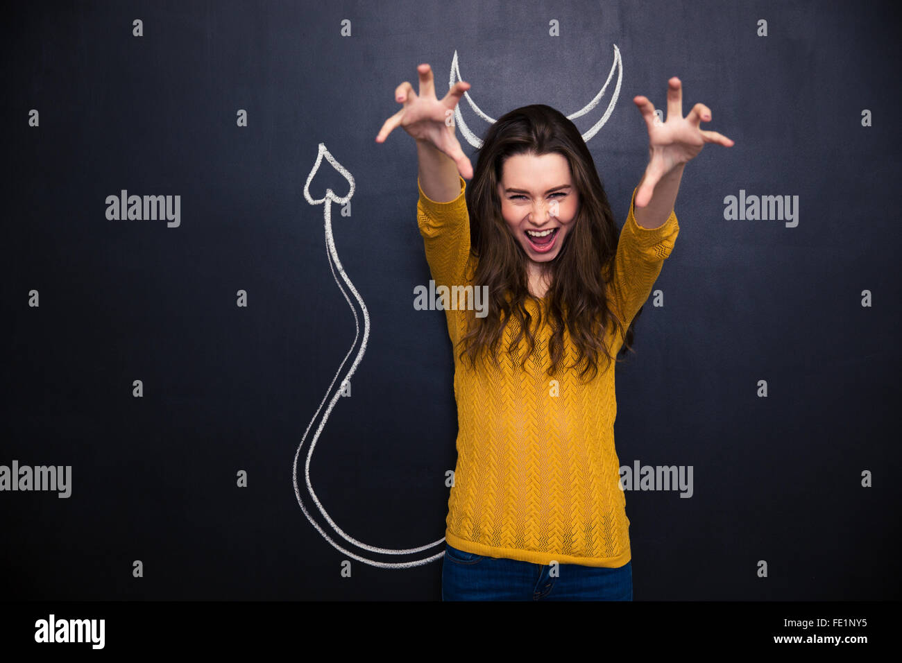 Shouting young woman with raised hands pretending devil standing over chalkboard background Stock Photo
