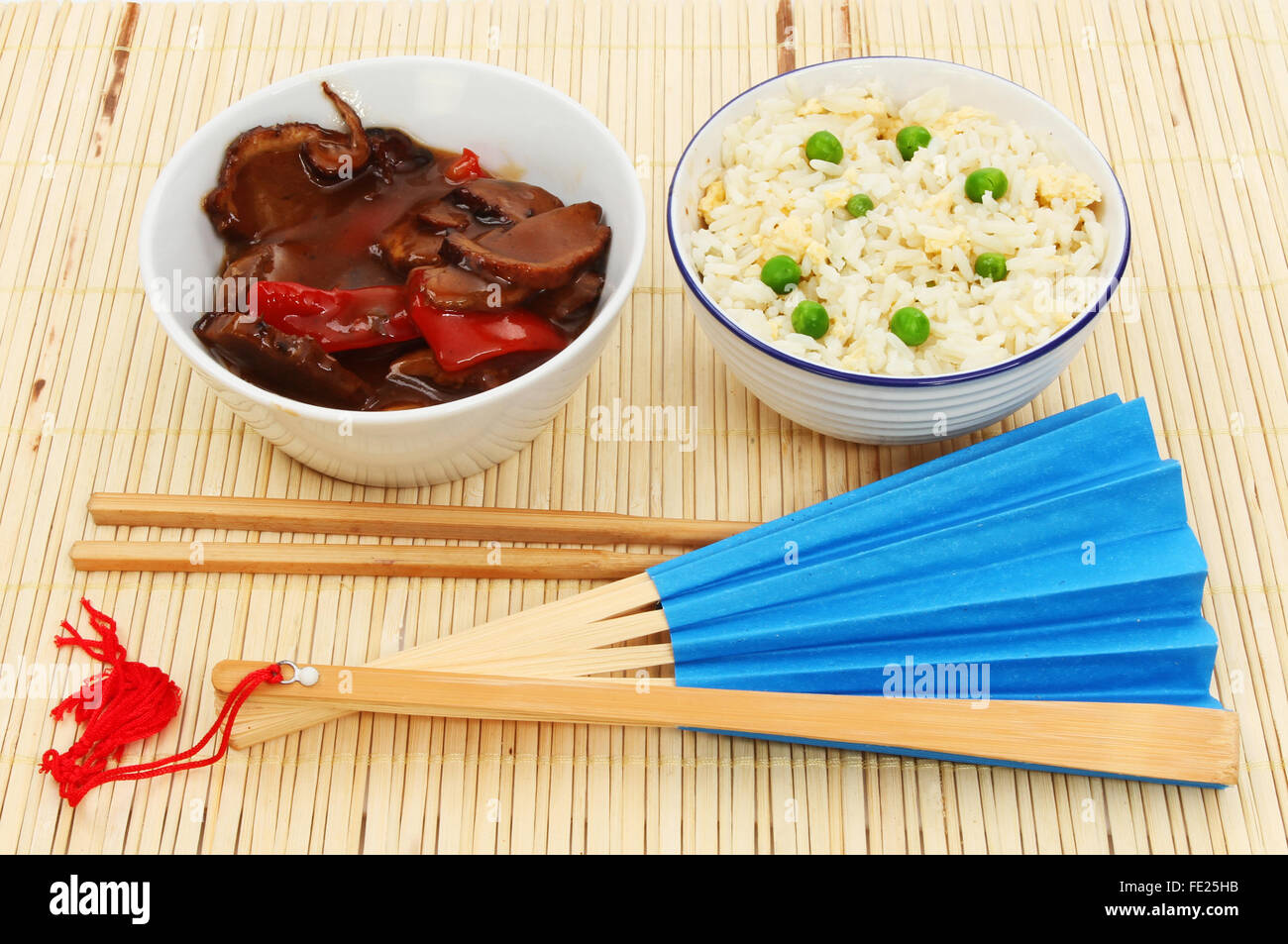 Oriental meal, egg fried rice and duck in plum sauce in bowls with chopsticks and a blue fan on a bamboo mat Stock Photo