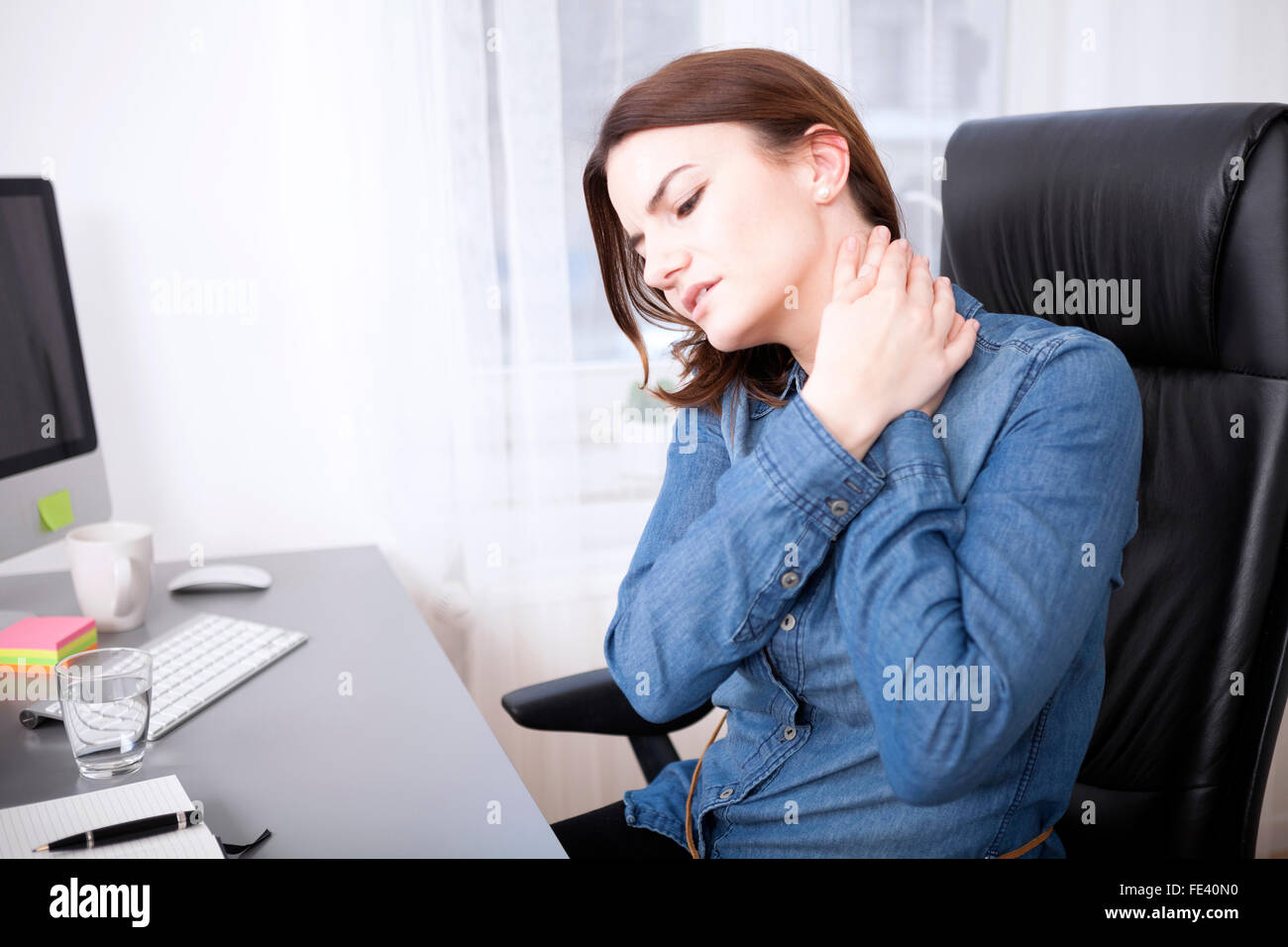 Close up Tired Young Office Girl in Denim Shirt Sitting at her Worktable While Holding her Neck with Both Hands Stock Photo