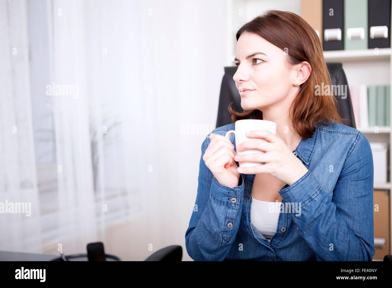 Businesswoman sitting thinking at her desk as she enjoys a coffee break ...