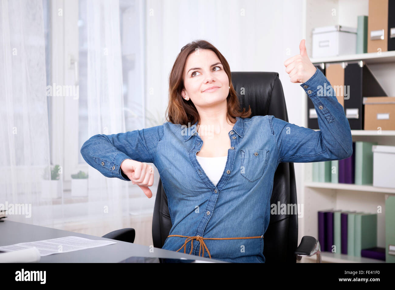 Close up Thoughtful Young Office Woman Sitting on her Chair Showing Thumbs Up and Down Hand Signs While Looking Up. Stock Photo