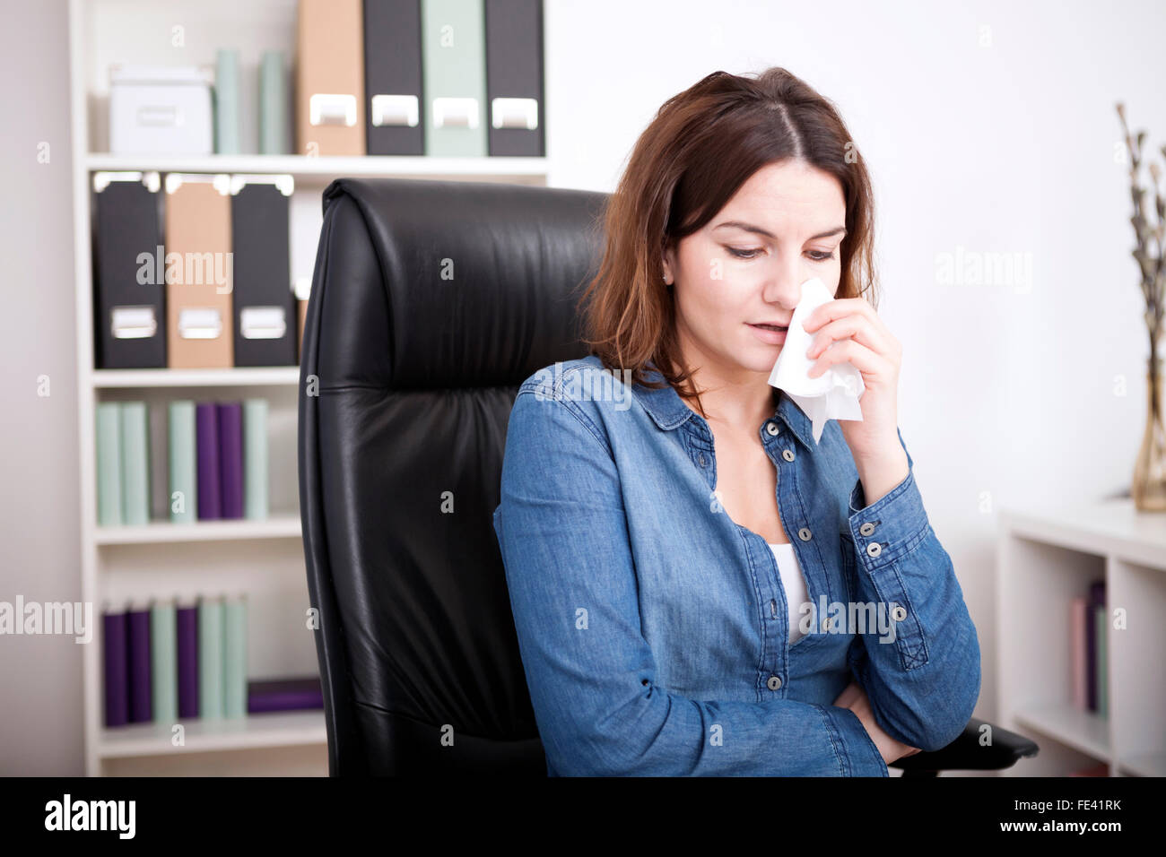 Distressed attractive businesswoman sitting crying in the office wiping her eye with a tissue due to bereavement of failure at w Stock Photo