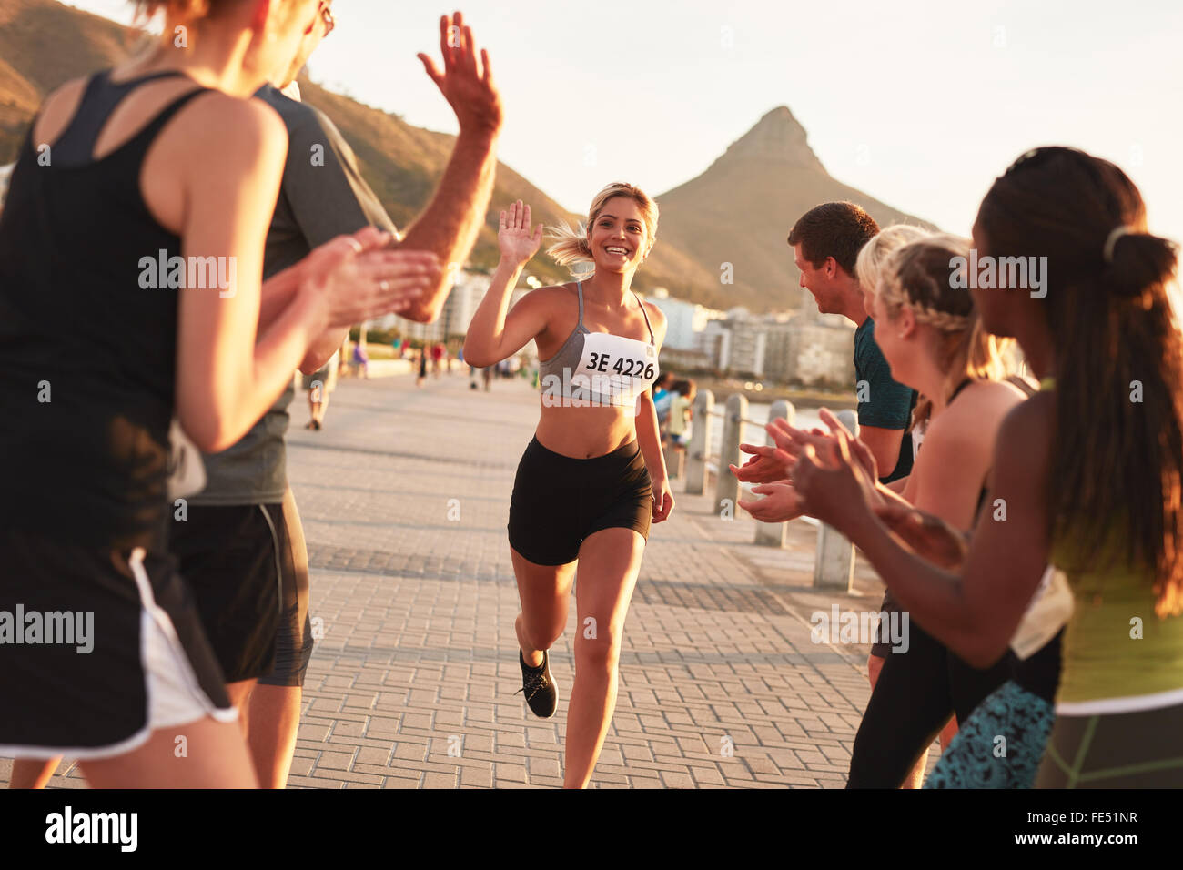 Group of spectators cheering runners just before the finish line. Female runner finishing the race with her team applauding her Stock Photo