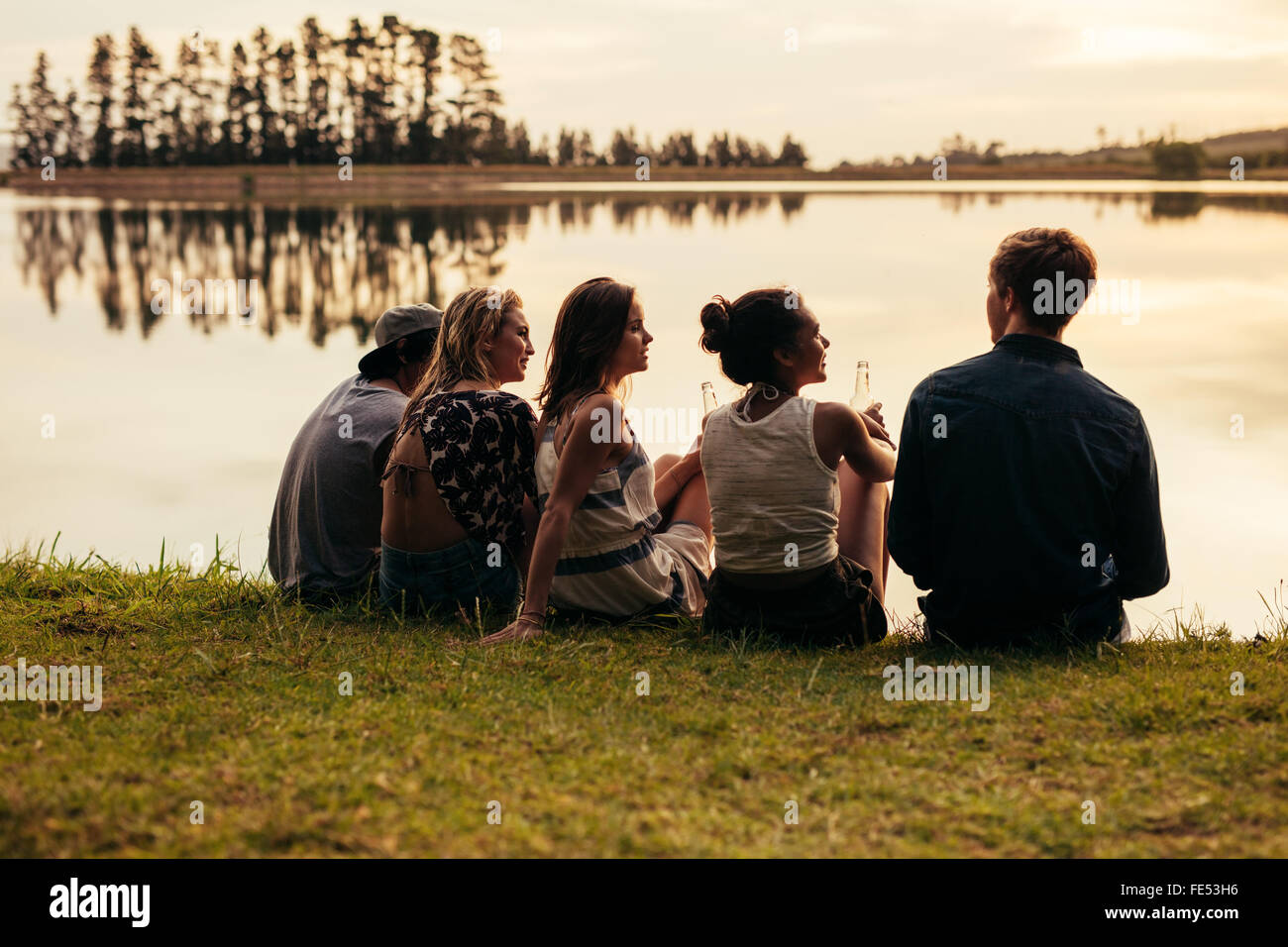 Rear view portrait of group of young friends relaxing by a lake. Young people sitting together by a lake. Stock Photo