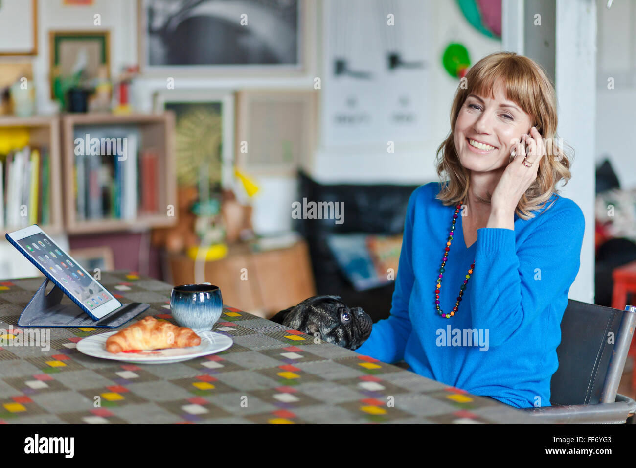 Woman smiles with a dog holding a phone at the living room table. Stock Photo