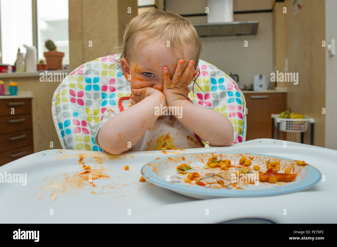 A baby girl (age 15 months) eats a meal of sweet corn and tomato sauce in a messy manner and plays with her food on a high chair Stock Photo