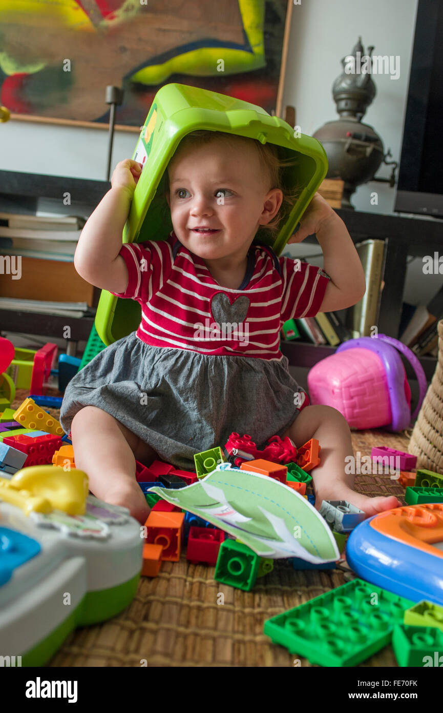 A baby girl (age 15 months) has fun putting a plastic box over her head as she plays with Lego and other toys in her living room Stock Photo