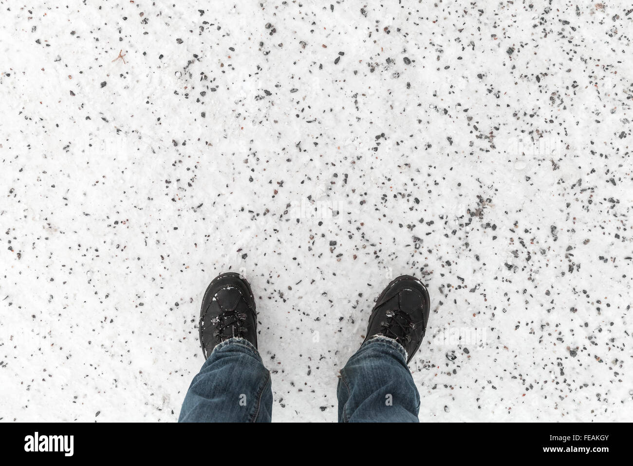 Male feet in blue jeans and black shoes standing on snowy winter road with anti-slip granite chippings, first person view Stock Photo