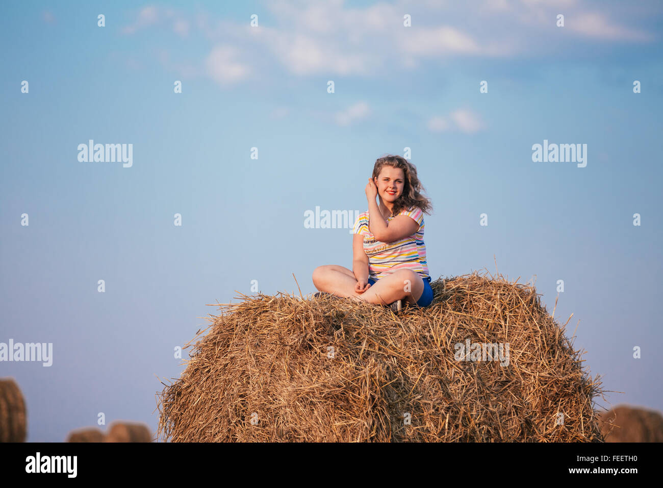 Beautiful smiling caucasian plus Size Young Woman Girl Sitting On Haystack on summer sunny field. Blue cloudy sky background. Stock Photo