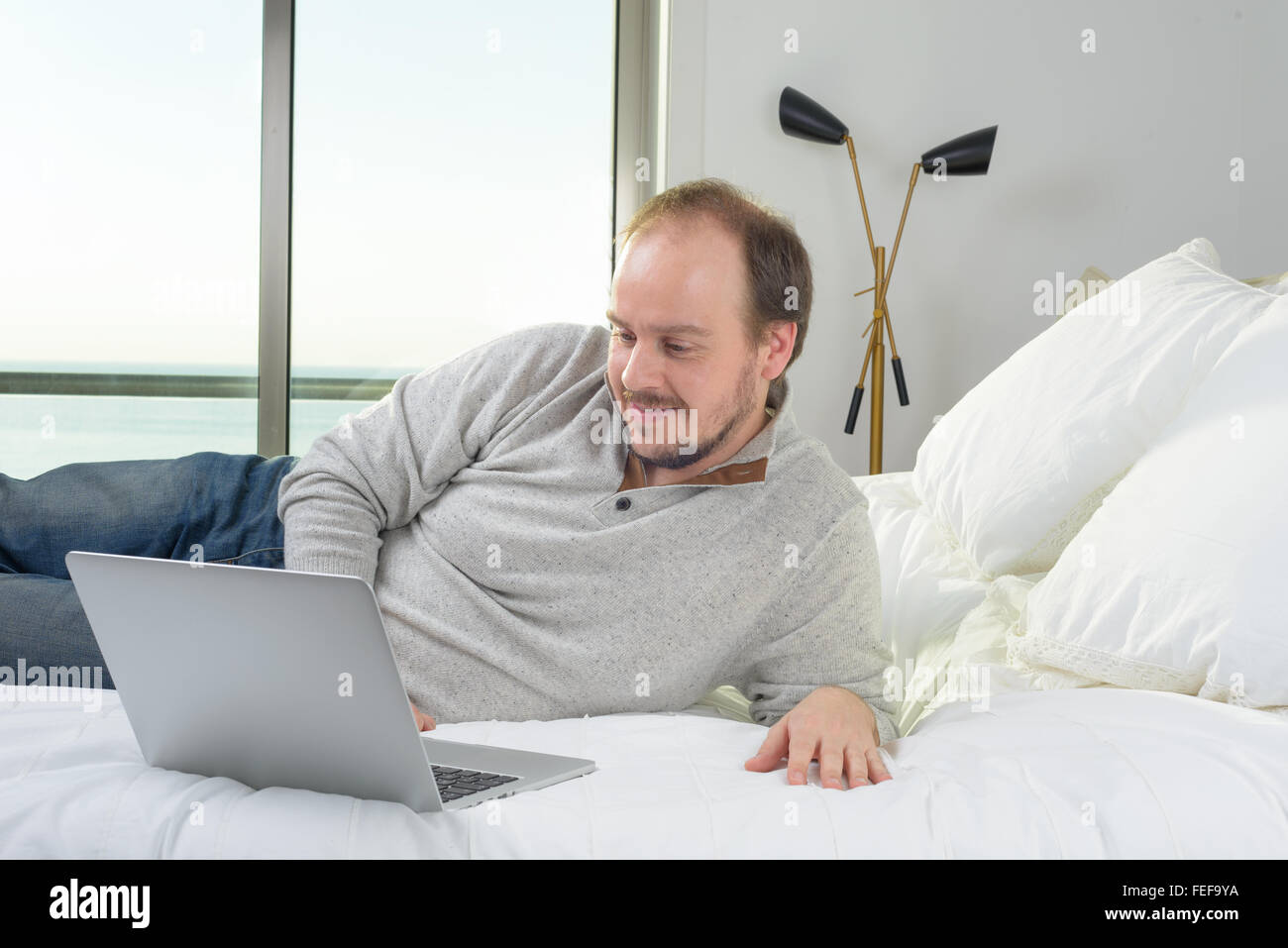 man lying in bed smiling working on laptop computer Stock Photo - Alamy