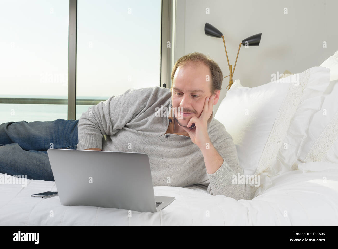 man lying on the bed smiling while working on laptop computer with ...
