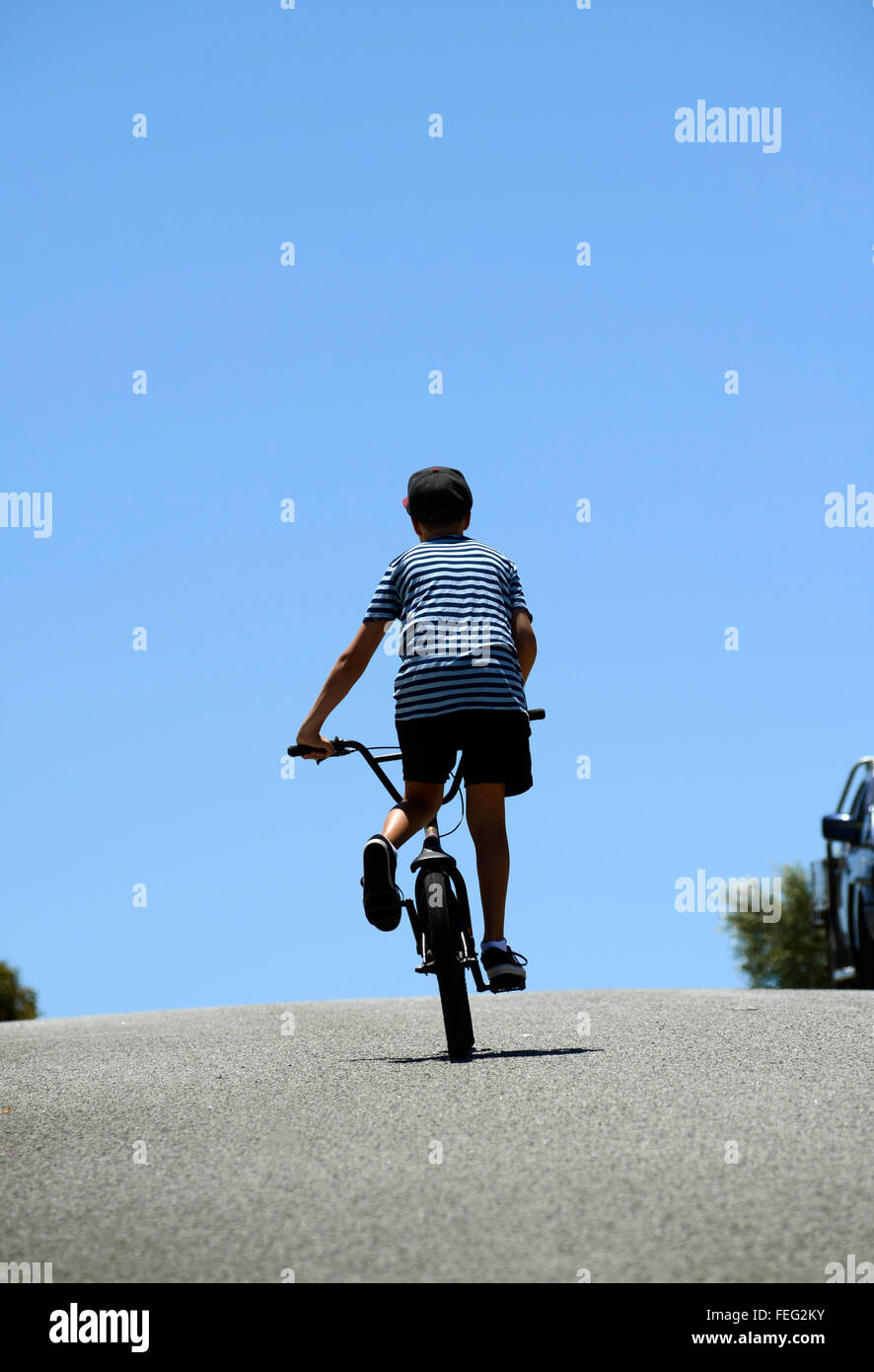 Boy with back to camera rides his BMX bicycle up a hill, almost silhouetted against a blue sky Stock Photo