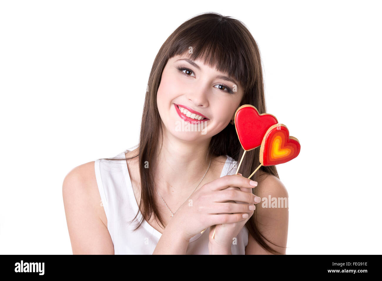 Headshot portrait of young beautiful cheerful woman holding two heart shaped red cookies, studio, white background, isolated Stock Photo