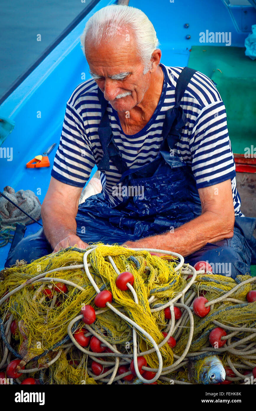 An older weather beaten fisherman wearing a nautical blue & white shirt  under overalls on boat works at finding fish in net Stock Photo - Alamy