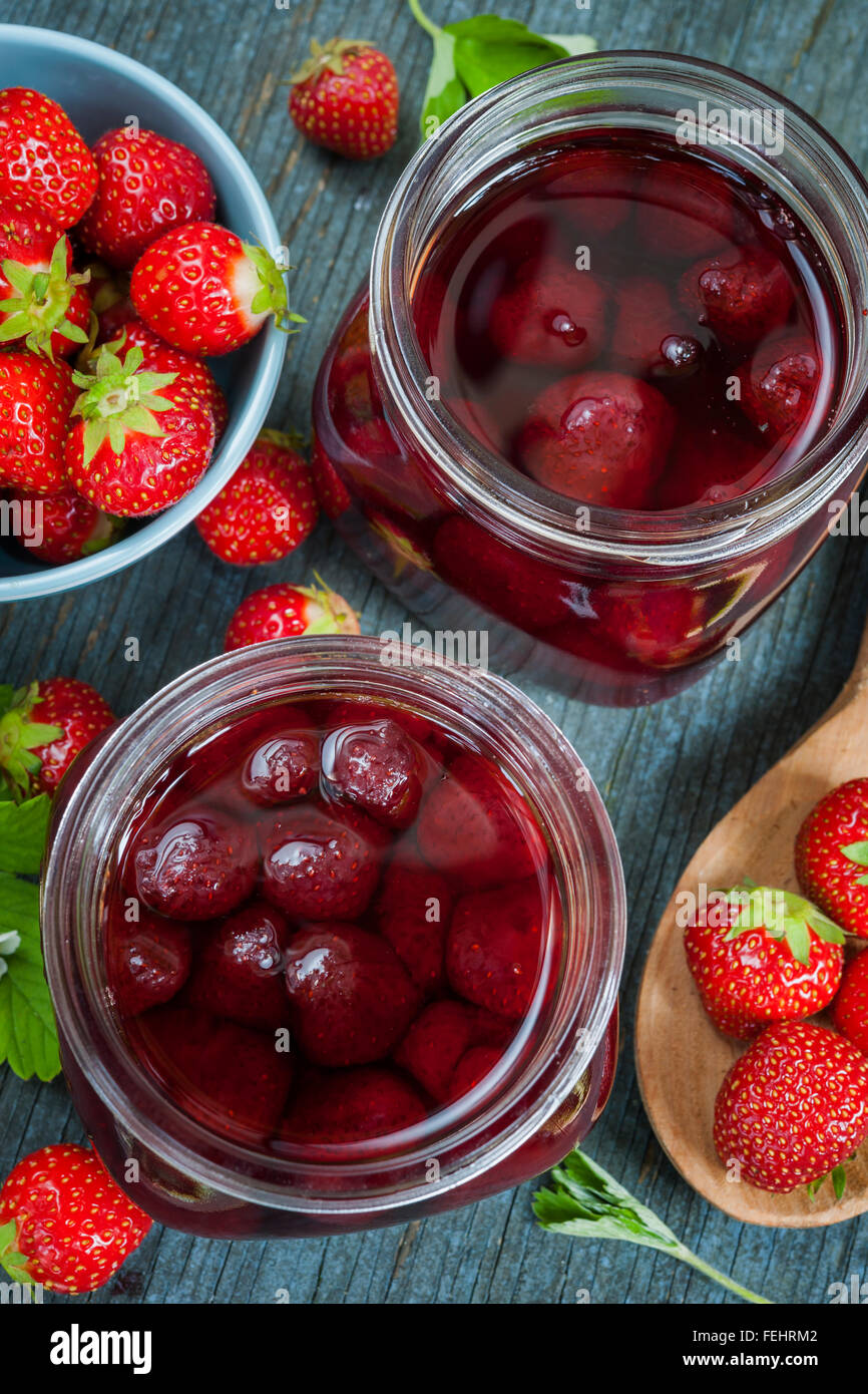 Glass jars with homemade strawberry preserve and fresh strawberries in bowl from above on blue rustic wood background, close up Stock Photo