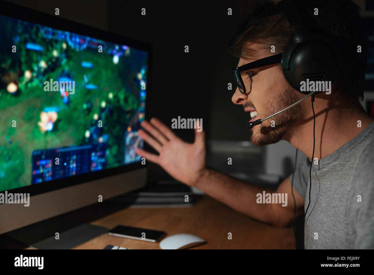 Cheerful young gamer in glasses playing game on computer using headphones Stock Photo
