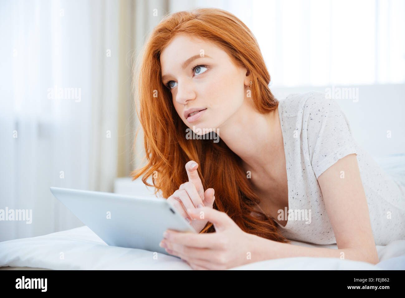 Redhead woman lying on the bed with tablet computer and thinking about something Stock Photo