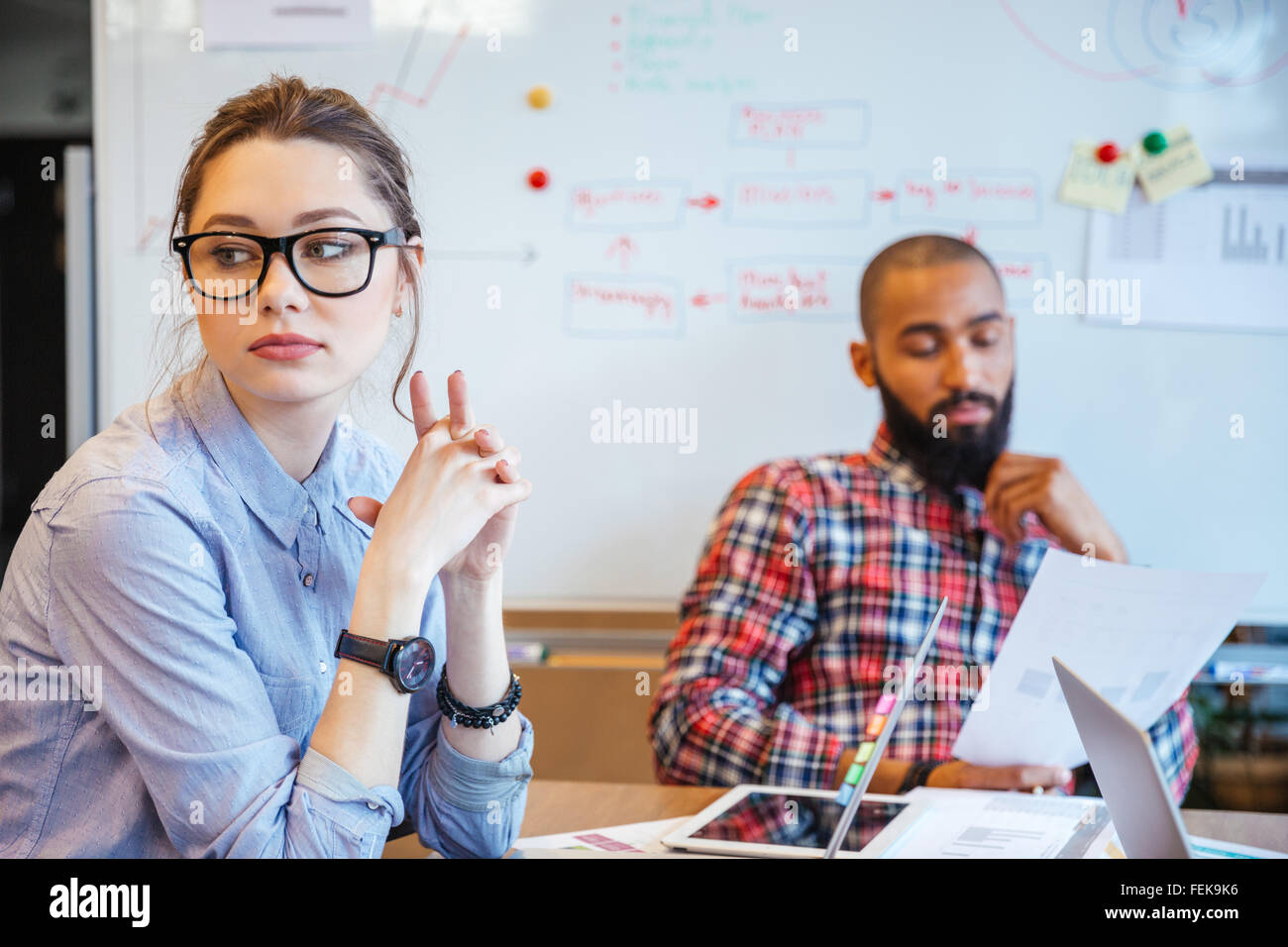 Pensive young woman in glasses sitting in conference room and thinking while her male colleague reading Stock Photo