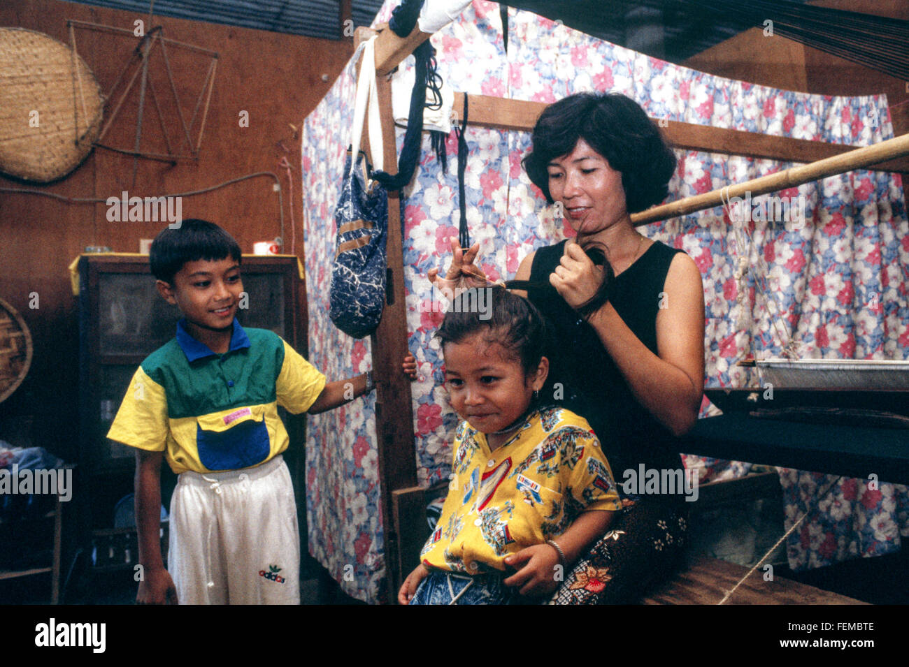 Young brother & sister get ready for school in their tiny makeshift home in Laos & their mother tidies the daughter's long hair. Stock Photo