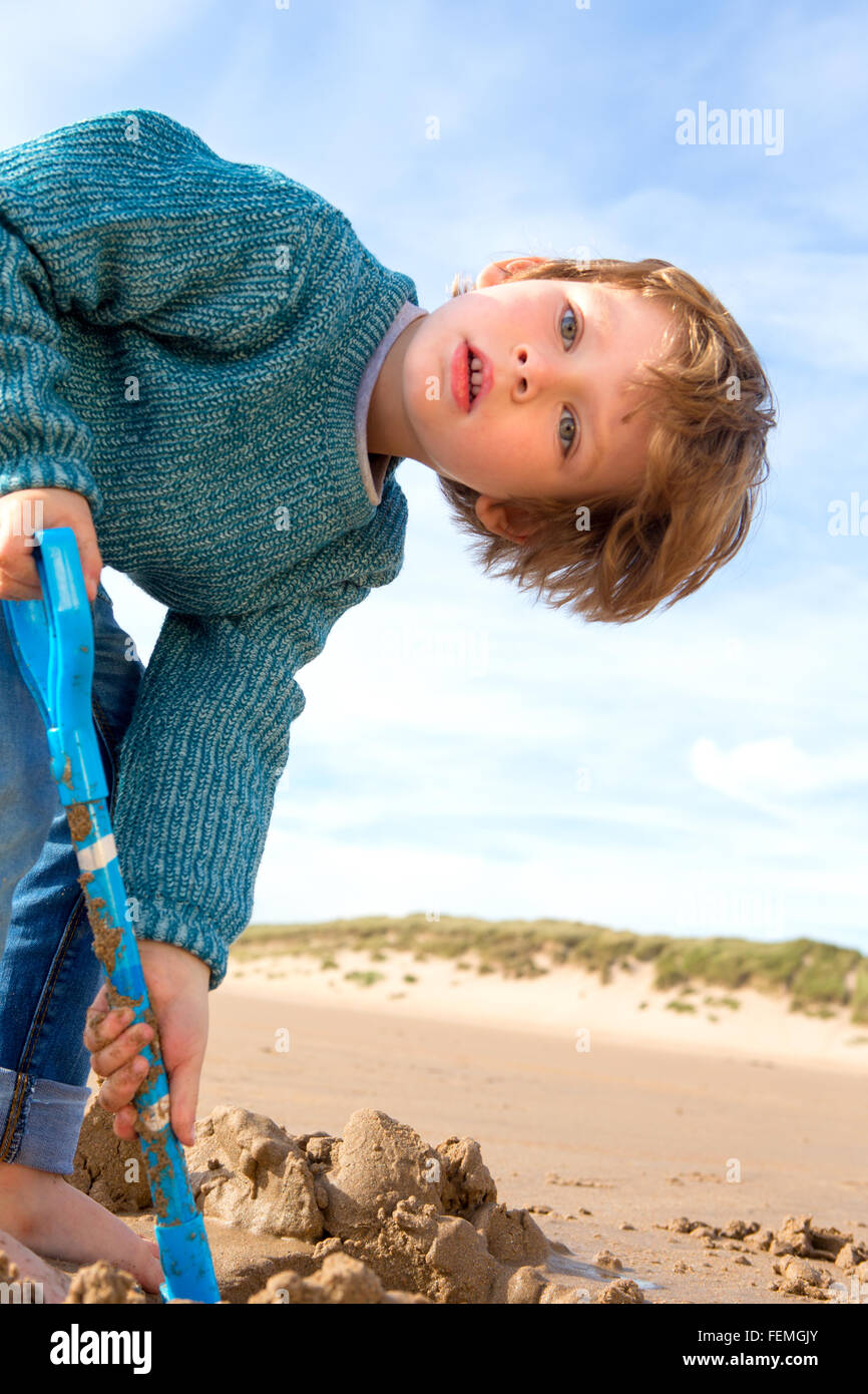 Young boy digging on the beach with a spade. He is bent over and ...