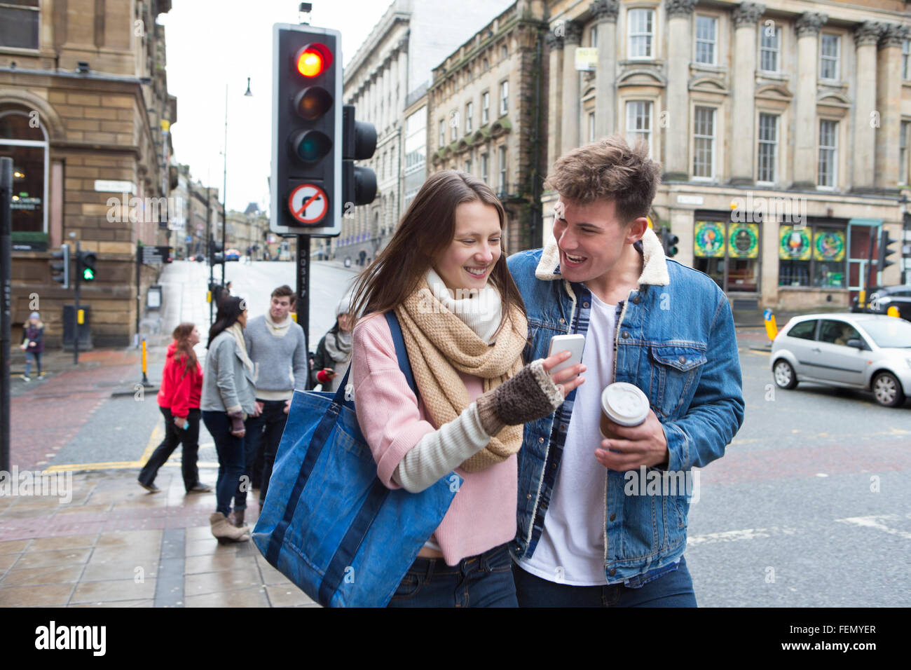 A young couple can be seen walking down a street looking at a smartphone together. Other young adults can be seen behind them. Stock Photo