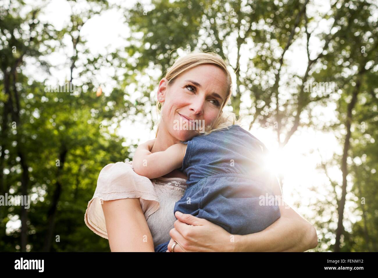 Mid adult woman carrying toddler daughter in sunlit park Stock Photo