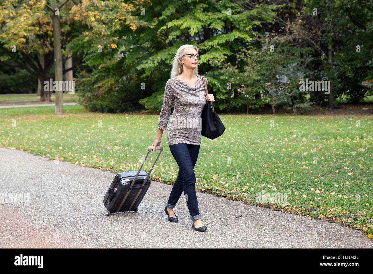 Mature woman walking through park pulling wheeled suitcase Stock Photo