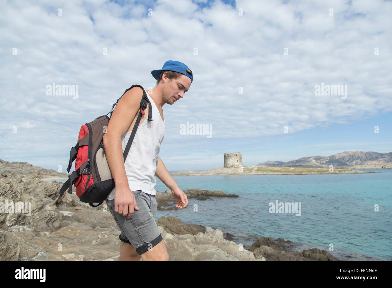 Young man on rocks by ocean carrying backpack looking down, Stintino