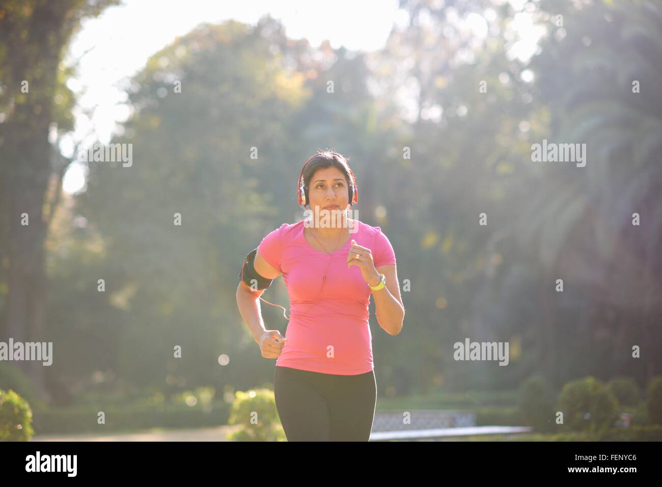 Front view of mature woman wearing headphones and armband jogging in park Stock Photo