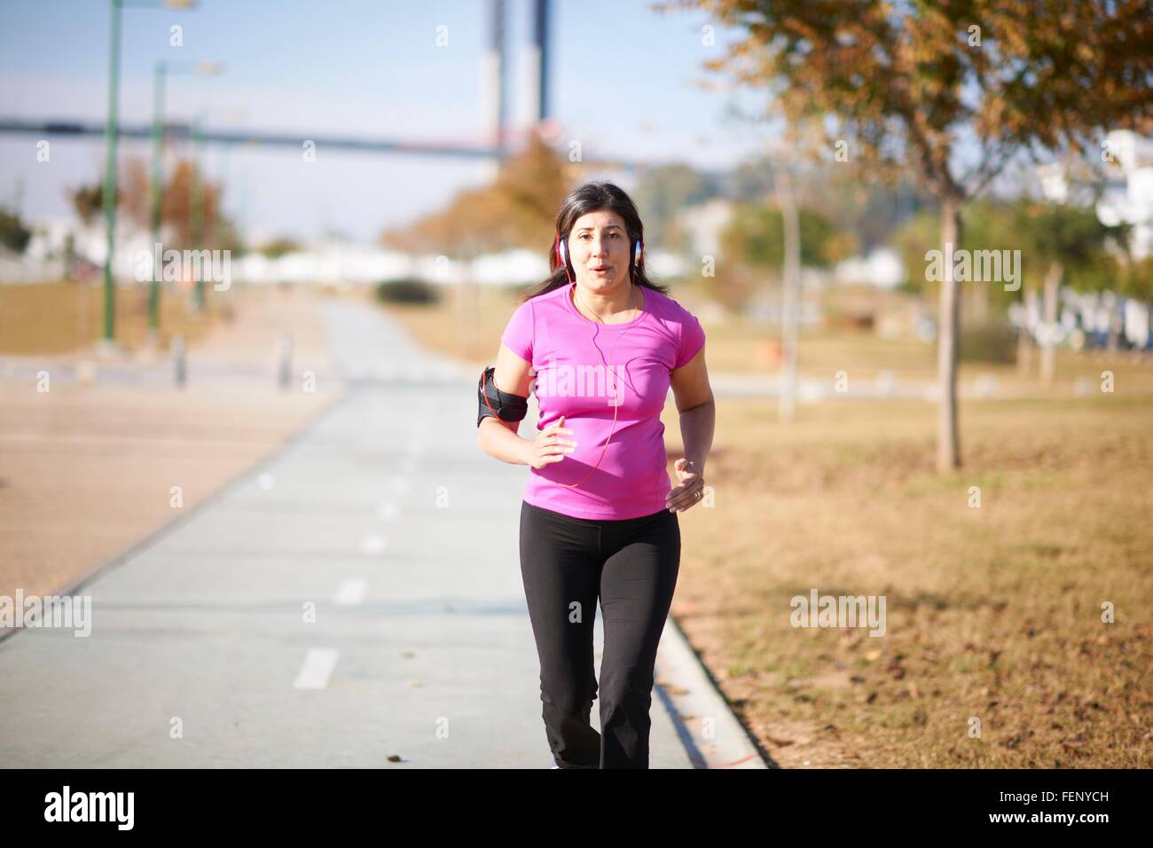 Front view of mature woman wearing headphones and armband jogging on pathway Stock Photo