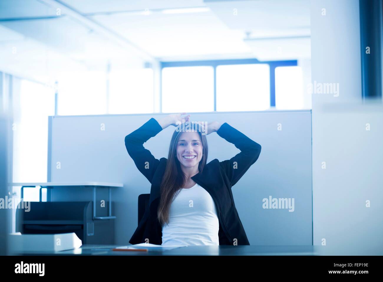 Young woman sitting at desk in office hands behind head looking at camera smiling Stock Photo