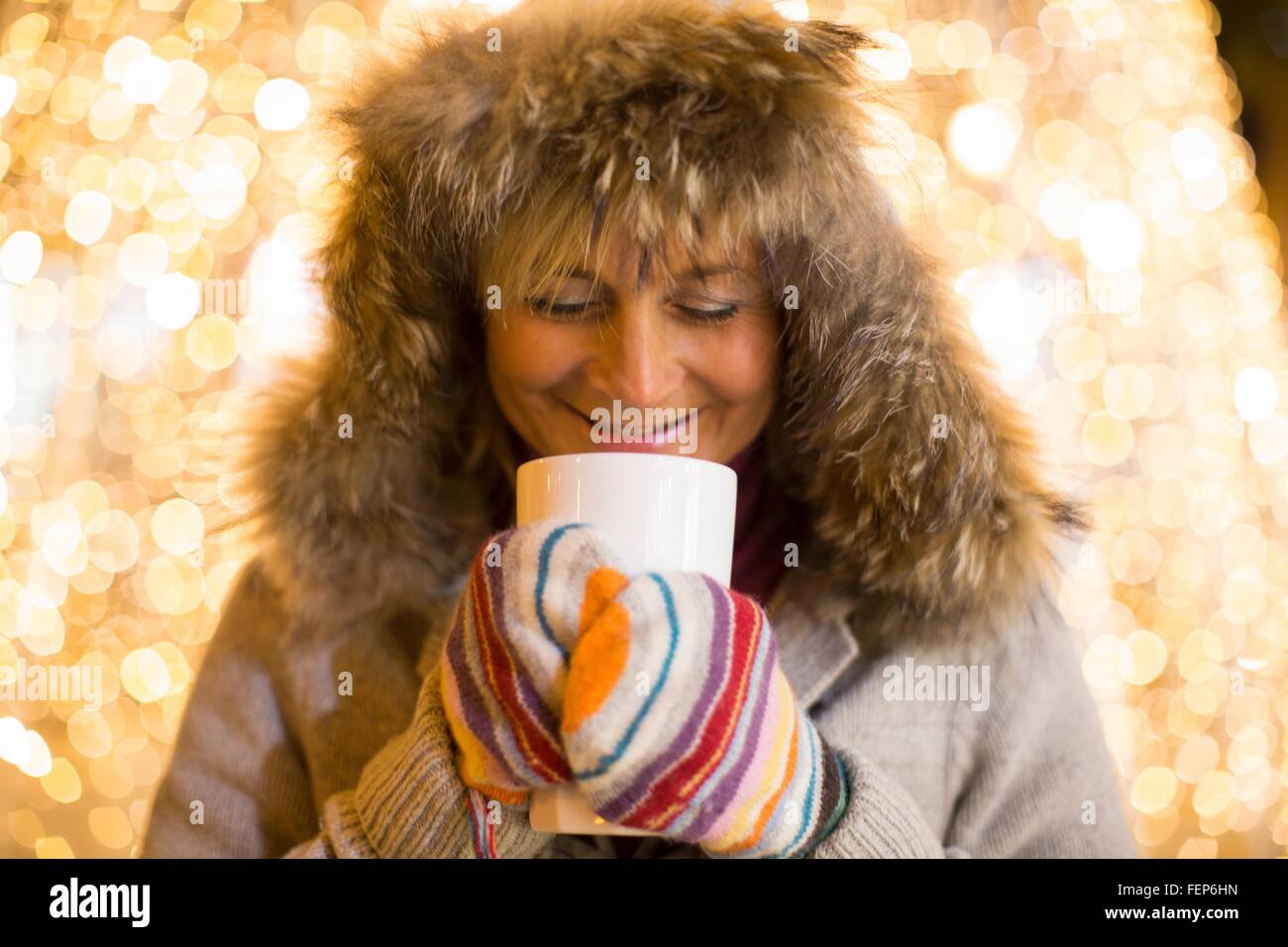 Mature woman wearing furry hood and mittens holding hot drink in front of xmas lights Stock Photo