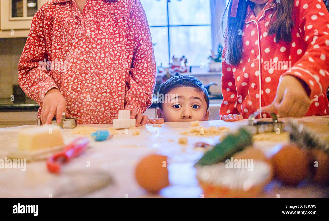 Boy in kitchen making cookies with sisters peeking over kitchen counter looking at camera Stock Photo