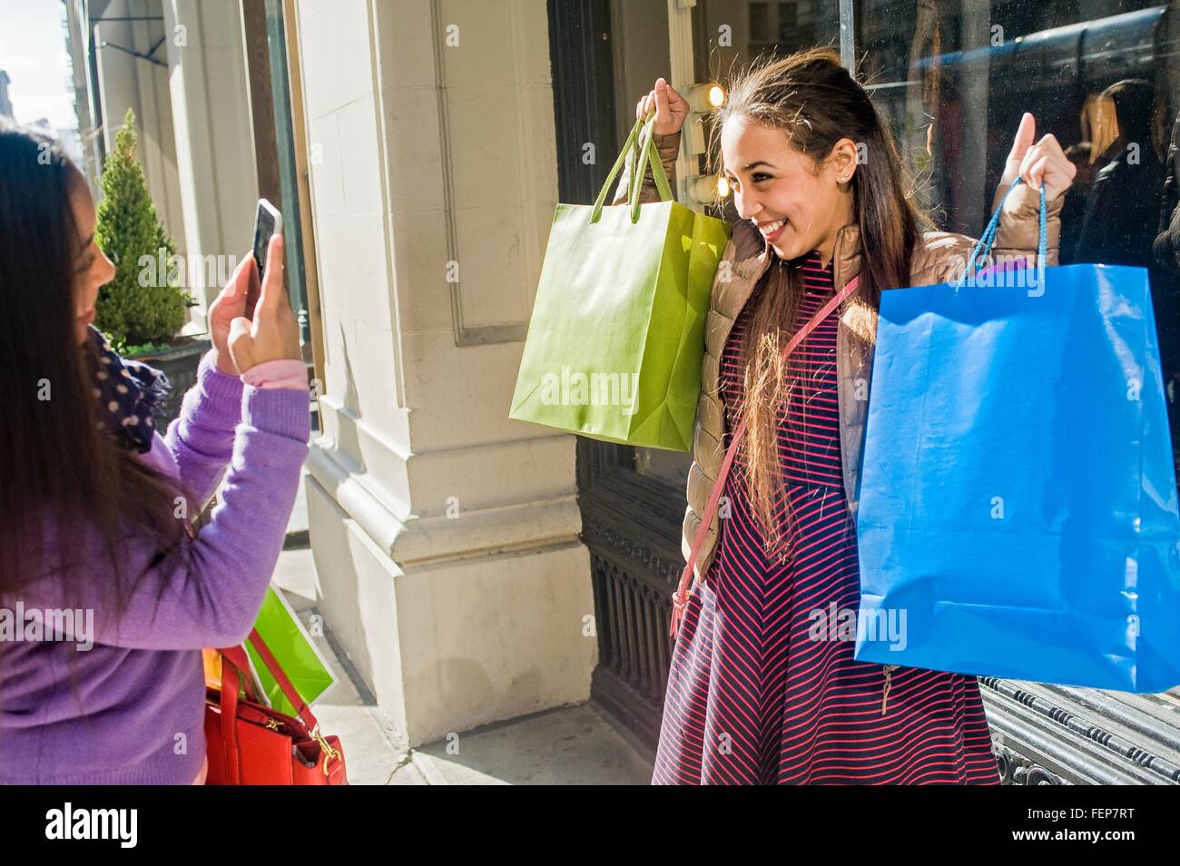 Young female adult twins in city photographing each other with shopping bags on smartphone Stock Photo