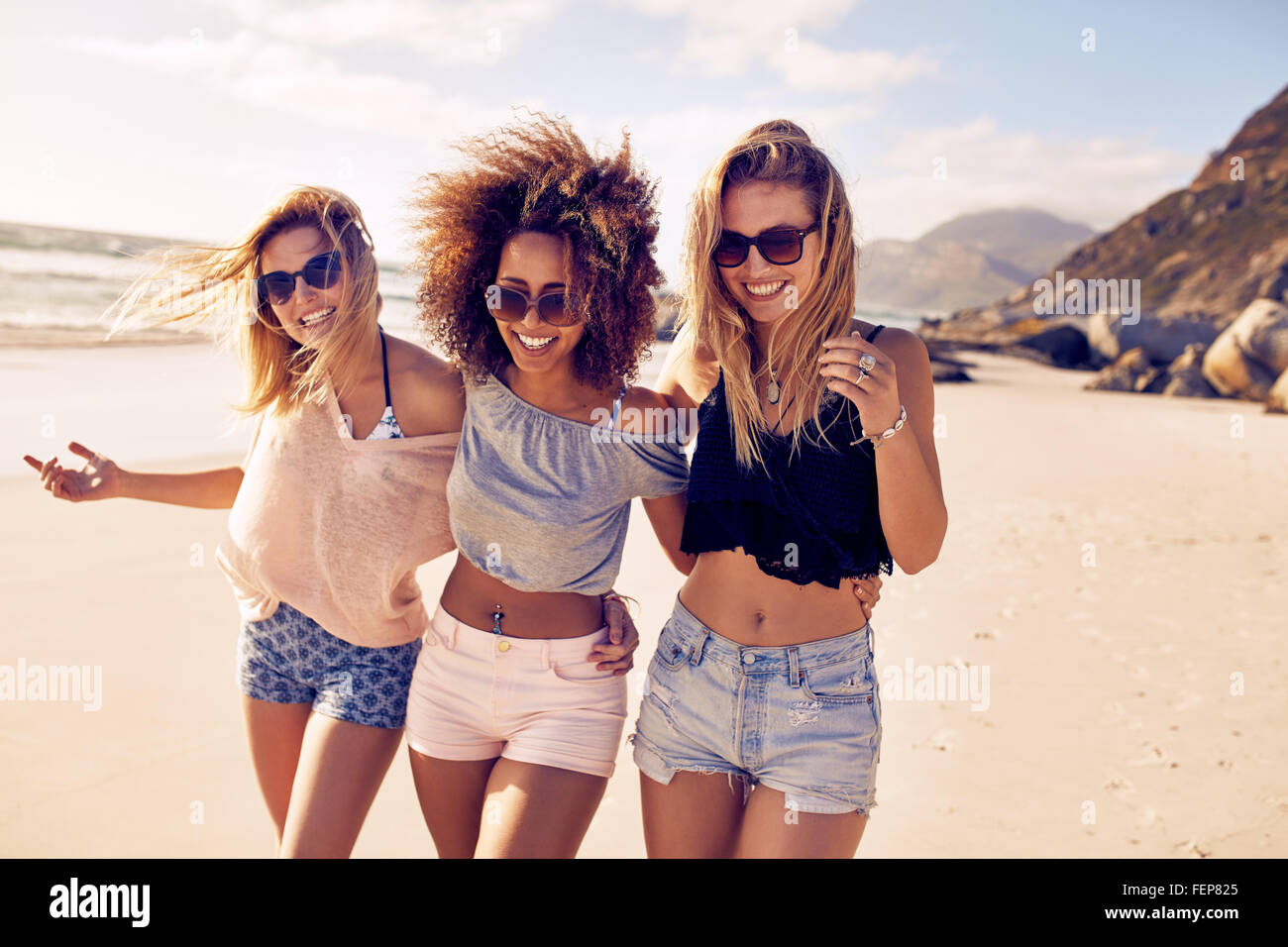 Portrait of three young female friends walking on the sea shore looking at camera laughing. Multiracial young women strolling al Stock Photo