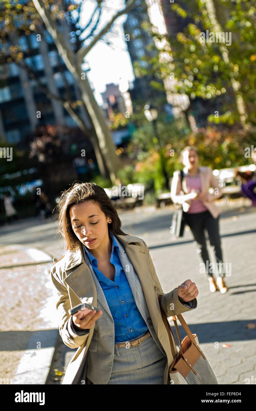 Young woman reading smartphone whilst walking through city park Stock Photo