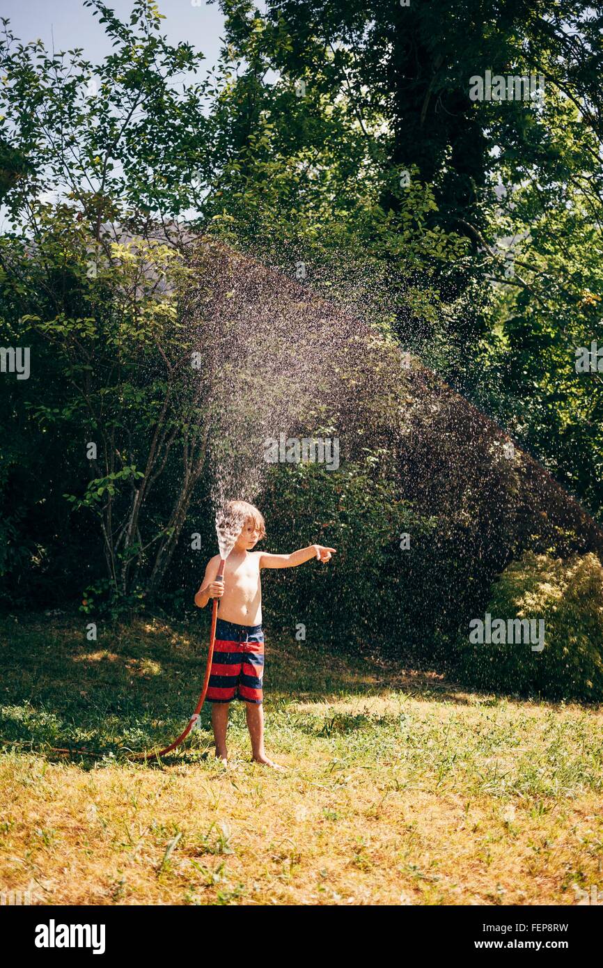 Full length view of boy in garden holding hosepipe spraying water, looking away pointing, Bludenz, Vorarlberg, Austria Stock Photo
