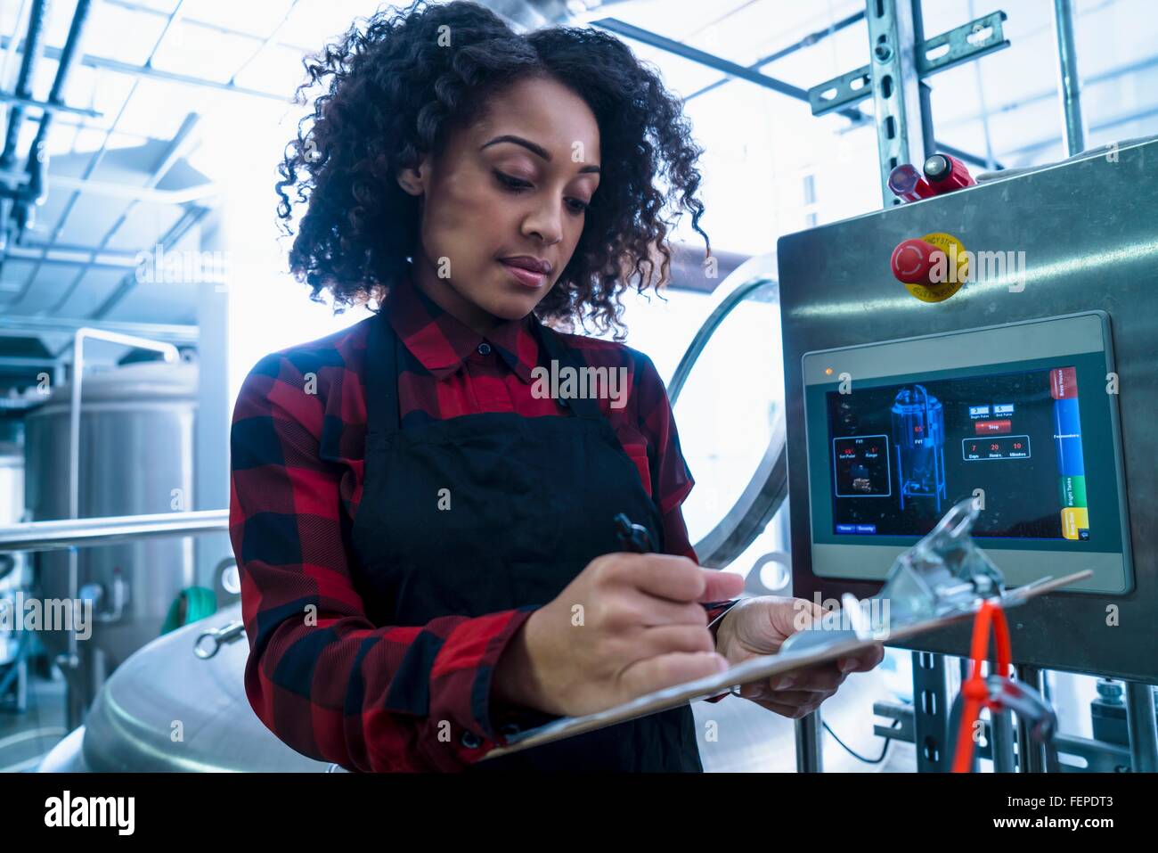 Mid adult woman in brewery monitoring computer equipment, looking down writing on clipboard Stock Photo