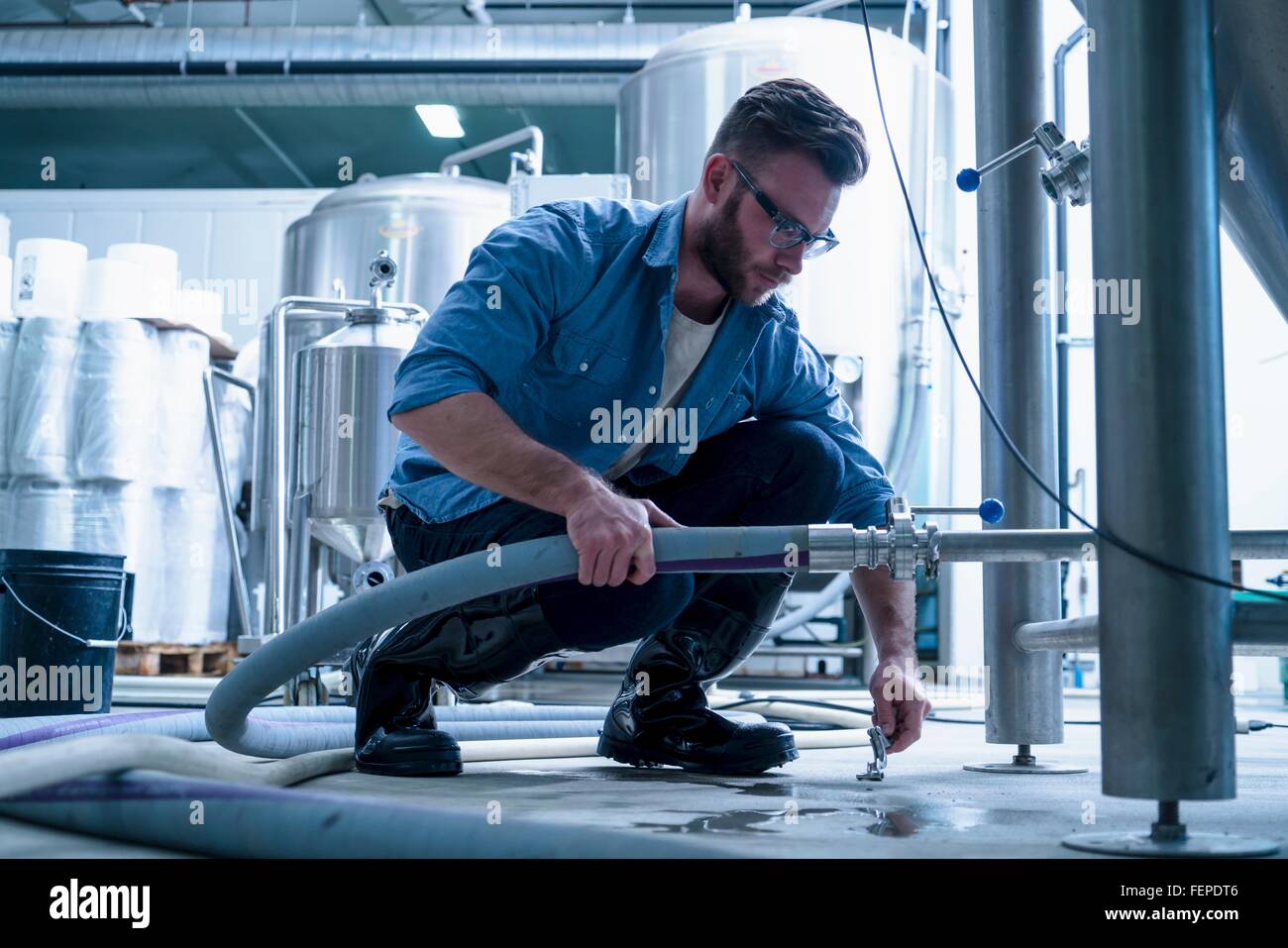 Young man in brewery crouching down connecting pipe Stock Photo