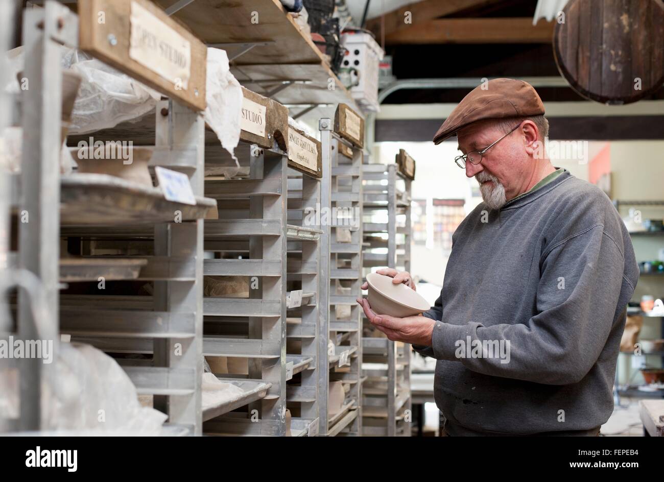 Potter wearing flat cap in storage room quality checking unfinished clay pots Stock Photo