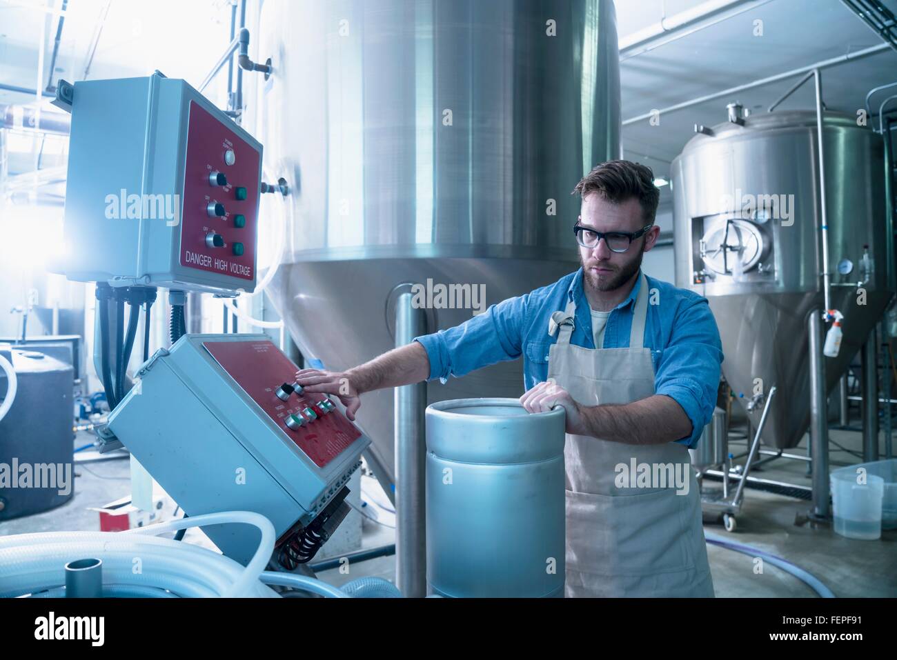 Young man in brewery by conical fermentation tank holding canister, pressing button on control panel Stock Photo