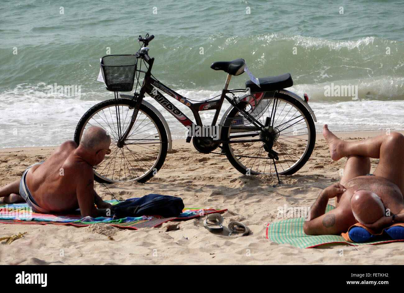 A Bicycle with a passenger pillion seat parked on the beach in Pattaya Thailand with two elderly males sunbathing and relaxing Stock Photo