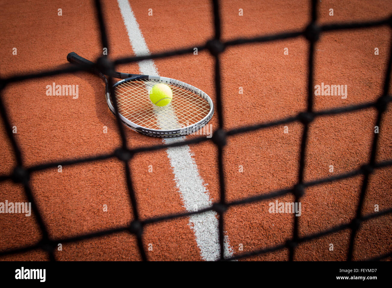 Tennis racket with a yellow ball on a brick red court through the net Stock Photo