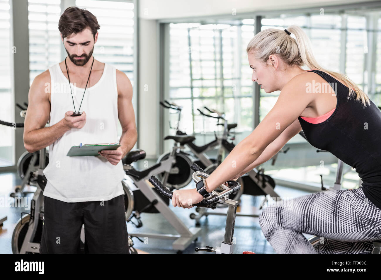 Trainer looking at stopwatch and woman using exercise bike Stock Photo